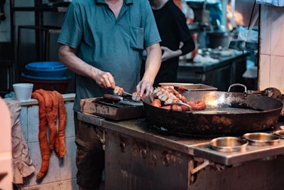 A person is preparing food in a kitchen setting, slicing pieces of meat or sausage on a cutting board next to a large frying pan. The setting appears to be a commercial kitchen or street food stall, with various utensils and containers around, including a few bowls and a large blue tub in the background.