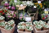 A vibrant display of various flowers arranged in bouquets at a market stall. The assortment includes tulips wrapped in paper, sunflowers, roses, and other colorful blossoms, with visible price tags indicating their cost.