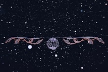 Strings of ornamental lights hang against a dark night sky, adorned with swirling and intricate designs. Snowflakes are falling softly, creating a serene and wintry atmosphere.