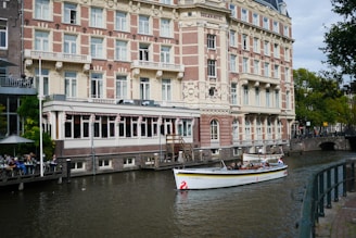 A boat is floating on a calm canal in front of a large, ornate, multi-story building labeled 'Doelen Hotel'. The canal is bordered by a sidewalk with a railing. On the left there is an outdoor seating area with people at tables under umbrellas. The boat has people on board and is adorned with decorative elements. The building has a mix of brick and stonework, with numerous windows and intricate architectural detailing. Trees are visible in the background.
