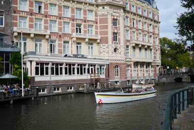 A boat is floating on a calm canal in front of a large, ornate, multi-story building labeled 'Doelen Hotel'. The canal is bordered by a sidewalk with a railing. On the left there is an outdoor seating area with people at tables under umbrellas. The boat has people on board and is adorned with decorative elements. The building has a mix of brick and stonework, with numerous windows and intricate architectural detailing. Trees are visible in the background.