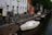 A canal lined with bicycles and a small sailboat moored against a brick wall. Residential buildings with large windows and a red-brick facade are visible in the background. Several cars and colorful canal boats are also present along the water's edge, with trees overhanging the street.