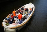 A group of people are sitting in a large open boat on the water, engaged in conversation and enjoying snacks. They appear to be participating in a guided boat tour or group outing. Several people are wearing bright orange shirts, and the boat is equipped with seating and a table filled with various items.