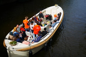 A group of people are sitting in a large open boat on the water, engaged in conversation and enjoying snacks. They appear to be participating in a guided boat tour or group outing. Several people are wearing bright orange shirts, and the boat is equipped with seating and a table filled with various items.
