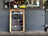 A restaurant exterior features a menu board with handwritten specials in various colors. The board lists dishes such as Linguine Italiana, Fettuccini Mataciana, Barramundi Fillet, and Beef Stroganoff with their respective prices. It leans against a gray brick wall, and part of the restaurant's name is visible through the window behind, alongside some seating and menus displayed inside.