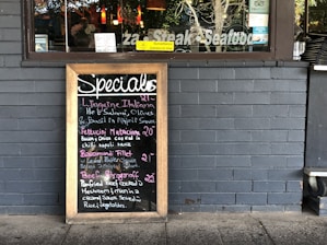 A restaurant exterior features a menu board with handwritten specials in various colors. The board lists dishes such as Linguine Italiana, Fettuccini Mataciana, Barramundi Fillet, and Beef Stroganoff with their respective prices. It leans against a gray brick wall, and part of the restaurant's name is visible through the window behind, alongside some seating and menus displayed inside.