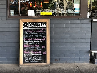 A restaurant exterior features a menu board with handwritten specials in various colors. The board lists dishes such as Linguine Italiana, Fettuccini Mataciana, Barramundi Fillet, and Beef Stroganoff with their respective prices. It leans against a gray brick wall, and part of the restaurant's name is visible through the window behind, alongside some seating and menus displayed inside.