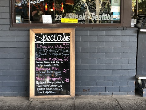 A restaurant exterior features a menu board with handwritten specials in various colors. The board lists dishes such as Linguine Italiana, Fettuccini Mataciana, Barramundi Fillet, and Beef Stroganoff with their respective prices. It leans against a gray brick wall, and part of the restaurant's name is visible through the window behind, alongside some seating and menus displayed inside.
