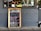 A restaurant exterior features a menu board with handwritten specials in various colors. The board lists dishes such as Linguine Italiana, Fettuccini Mataciana, Barramundi Fillet, and Beef Stroganoff with their respective prices. It leans against a gray brick wall, and part of the restaurant's name is visible through the window behind, alongside some seating and menus displayed inside.