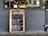 A restaurant exterior features a menu board with handwritten specials in various colors. The board lists dishes such as Linguine Italiana, Fettuccini Mataciana, Barramundi Fillet, and Beef Stroganoff with their respective prices. It leans against a gray brick wall, and part of the restaurant's name is visible through the window behind, alongside some seating and menus displayed inside.