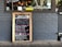 A restaurant exterior features a menu board with handwritten specials in various colors. The board lists dishes such as Linguine Italiana, Fettuccini Mataciana, Barramundi Fillet, and Beef Stroganoff with their respective prices. It leans against a gray brick wall, and part of the restaurant's name is visible through the window behind, alongside some seating and menus displayed inside.