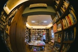 A cozy, warmly lit bookstore filled with numerous shelves of books lining the walls. A person wearing a red beanie stands reading or browsing. The perspective is slightly distorted as though taken with a fisheye lens, emphasizing the abundance of books and creating a snug, enclosed atmosphere.