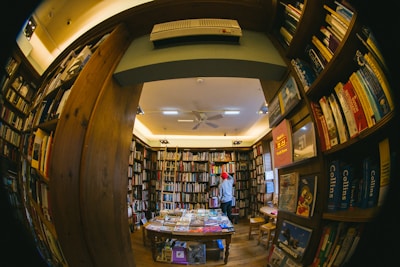 A cozy, warmly lit bookstore filled with numerous shelves of books lining the walls. A person wearing a red beanie stands reading or browsing. The perspective is slightly distorted as though taken with a fisheye lens, emphasizing the abundance of books and creating a snug, enclosed atmosphere.