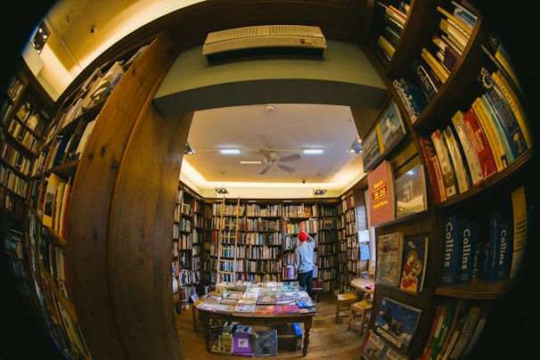 A cozy, warmly lit bookstore filled with numerous shelves of books lining the walls. A person wearing a red beanie stands reading or browsing. The perspective is slightly distorted as though taken with a fisheye lens, emphasizing the abundance of books and creating a snug, enclosed atmosphere.