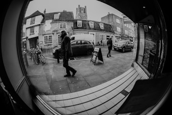 A black and white street view captured through a fisheye lens, featuring two people walking on the sidewalk. The scene includes old brick buildings with storefronts, a parked car, bicycles on the left, and a signboard in the center. The reflective glass creates a warped, circular effect, adding a unique perspective.