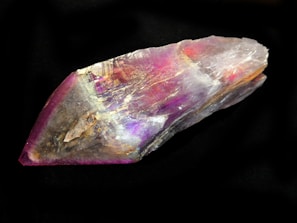 A strikingly beautiful crystal featuring a vibrant array of colors, including purple, pink, and hints of orange, set against a dark black background. The crystal has a rough, angular shape with visible striations and a reflective surface, creating a mesmerizing play of light and color.