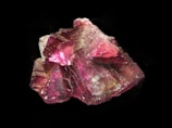 A rough mineral specimen with a rich, deep pink hue and an intricate crystalline structure. The surface reflects light, creating areas of intense color and sections with a translucent appearance. The mineral's facets and texture add complexity to its overall shape, set against a neutral dark background.