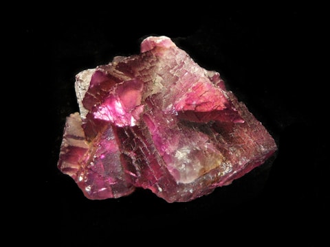 A rough mineral specimen with a rich, deep pink hue and an intricate crystalline structure. The surface reflects light, creating areas of intense color and sections with a translucent appearance. The mineral's facets and texture add complexity to its overall shape, set against a neutral dark background.