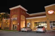 A shopping plaza at night features a store with a large sign reading 'Perfume & Sunglasses'. Two cars are parked in front of the building, and there are illuminated signs for other businesses. The architecture includes arched doorways and windows, with a prominent red-brown facade and a yellowish-tan building next to it.