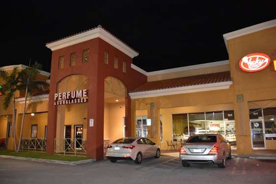 A shopping plaza at night features a store with a large sign reading 'Perfume & Sunglasses'. Two cars are parked in front of the building, and there are illuminated signs for other businesses. The architecture includes arched doorways and windows, with a prominent red-brown facade and a yellowish-tan building next to it.