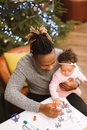 A person is holding a baby while coloring a page at a table with art supplies. A Christmas tree adorned with lights is visible in the background, creating a festive atmosphere.