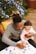 A person is holding a baby while coloring a page at a table with art supplies. A Christmas tree adorned with lights is visible in the background, creating a festive atmosphere.