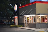A fast food restaurant with a brightly lit sign displaying the logo. The building features large glass windows and a modern exterior design. It is evening, and the lights from inside add warmth to the scene, contrasting with the darkening sky.