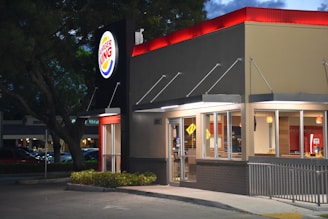 A fast food restaurant with a brightly lit sign displaying the logo. The building features large glass windows and a modern exterior design. It is evening, and the lights from inside add warmth to the scene, contrasting with the darkening sky.