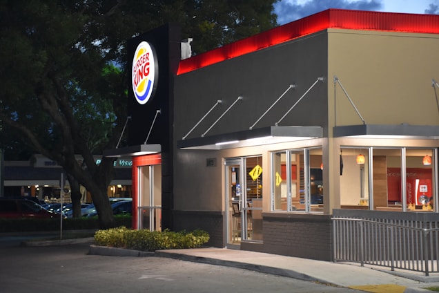 A fast food restaurant with a brightly lit sign displaying the logo. The building features large glass windows and a modern exterior design. It is evening, and the lights from inside add warmth to the scene, contrasting with the darkening sky.