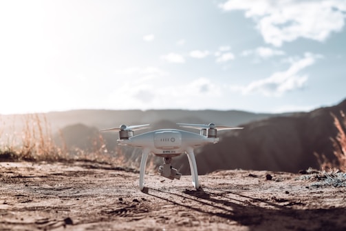 A white drone is positioned on a dirt path in a natural, outdoor setting. Surrounded by rolling hills and grass with a bright sky, the drone is equipped with a camera and seems ready for flight.