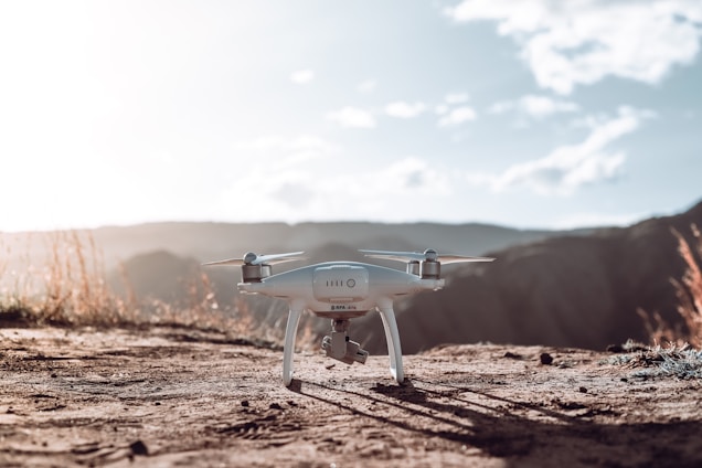 A white drone is positioned on a dirt path in a natural, outdoor setting. Surrounded by rolling hills and grass with a bright sky, the drone is equipped with a camera and seems ready for flight.