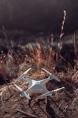 A white quadcopter drone rests on a patch of dry, brown grass and dirt. The background is softly blurred, suggesting an outdoor, natural setting.