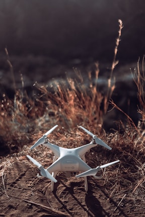 A white quadcopter drone rests on a patch of dry, brown grass and dirt. The background is softly blurred, suggesting an outdoor, natural setting.