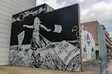 A striking black and white mural features a girl in a dress facing away, standing in tall grass. She holds a paper with inscriptions that read 'Words are lost' and 'Our future is unwritten.' The artwork is detailed, with a night sky and moon in the background. Words such as 'The sun fades' appear in the sky.