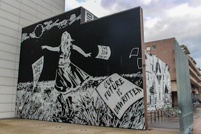 A striking black and white mural features a girl in a dress facing away, standing in tall grass. She holds a paper with inscriptions that read 'Words are lost' and 'Our future is unwritten.' The artwork is detailed, with a night sky and moon in the background. Words such as 'The sun fades' appear in the sky.