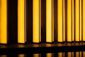Tall, vertical yellow light panels are arranged in a row, casting a warm glow and creating an abstract visual effect. The panels are reflected on the glossy surface below, adding depth to the scene.