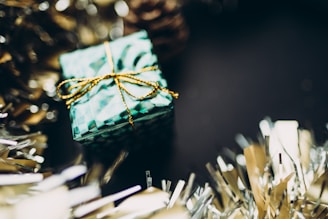 A small gift box wrapped in festive green patterned paper with a shiny gold ribbon sits amidst blurred tinsel decorations. The background is dark, creating a contrast that highlights the box and tinsel.