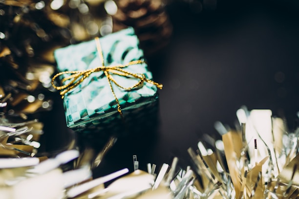 A small gift box wrapped in festive green patterned paper with a shiny gold ribbon sits amidst blurred tinsel decorations. The background is dark, creating a contrast that highlights the box and tinsel.