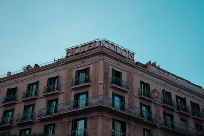A multi-story hotel building with classical architecture, featuring ornate balconies and a rooftop sign reading 'Hotel Colon'. The building has a warm beige facade with dark window shutters, contrasting against a clear blue sky.
