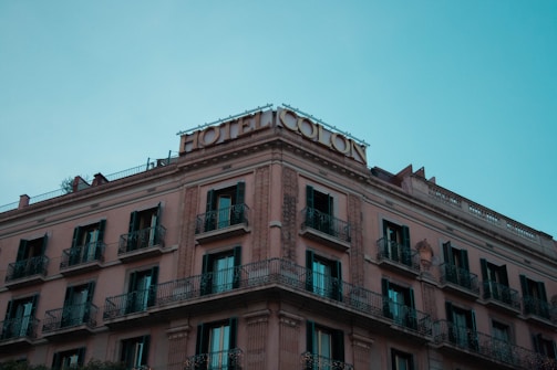 A multi-story hotel building with classical architecture, featuring ornate balconies and a rooftop sign reading 'Hotel Colon'. The building has a warm beige facade with dark window shutters, contrasting against a clear blue sky.