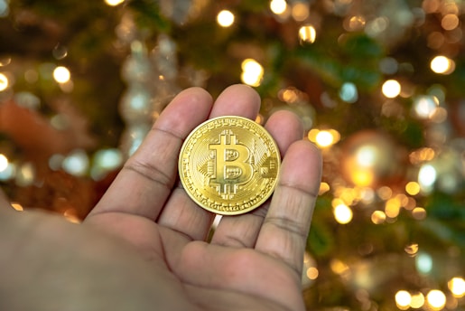 A close-up of a hand holding a gold-colored coin with the Bitcoin symbol on it. In the background, there are blurred lights creating a bokeh effect, giving a festive and warm ambiance.