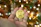 A close-up of a hand holding a gold-colored coin with the Bitcoin symbol on it. In the background, there are blurred lights creating a bokeh effect, giving a festive and warm ambiance.
