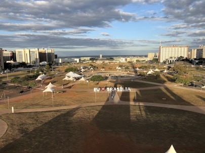 A grassy park with scattered white tents, surrounded by tall buildings. There is a large sign in the center displaying 'Eu ❤️ Brasília'. The sky is partly cloudy with scattered patches of sunlight casting long shadows on the ground.