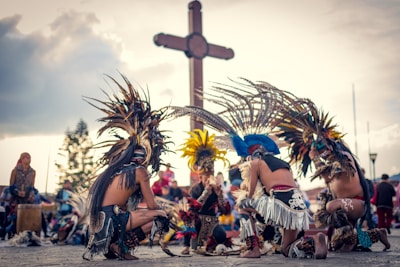 A group of people dressed in traditional, colorful feathered costumes, performing a cultural dance in an outdoor setting. In the background, a large wooden cross is visible against a partly cloudy sky.