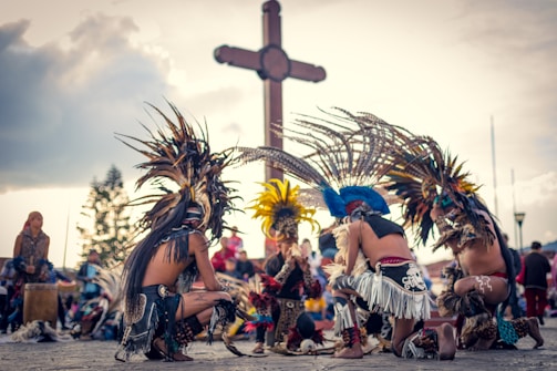 A group of people dressed in traditional, colorful feathered costumes, performing a cultural dance in an outdoor setting. In the background, a large wooden cross is visible against a partly cloudy sky.