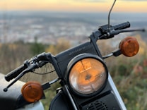 A close-up of a motorcycle's front headlight with two orange indicators on either side, showcasing the handlebars and a slightly blurred background of a landscape at dusk.