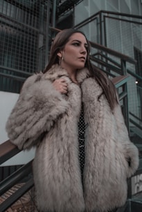 A person wearing a luxurious fur coat stands thoughtfully next to a modern, industrial staircase. The setting features metal railings and a mix of concrete and glass materials, adding a contemporary urban feel to the scene.