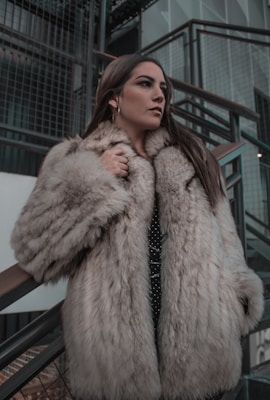 A person wearing a luxurious fur coat stands thoughtfully next to a modern, industrial staircase. The setting features metal railings and a mix of concrete and glass materials, adding a contemporary urban feel to the scene.