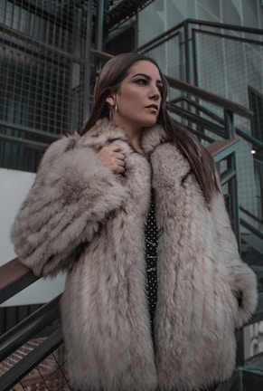 A person wearing a luxurious fur coat stands thoughtfully next to a modern, industrial staircase. The setting features metal railings and a mix of concrete and glass materials, adding a contemporary urban feel to the scene.