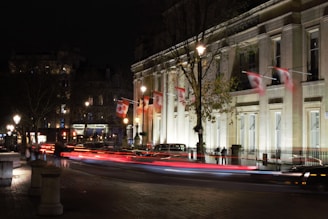 A nighttime city scene featuring a well-lit building adorned with Canadian flags. The street is busy with motion blur from passing vehicles, creating streaks of light. Trees and lampposts line the road, and a sense of urban life is evident.